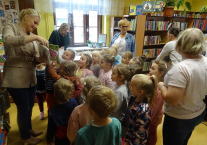 p. Dyrektor Miejsko- Gminnej Biblioteki Publicznej w Kleczewie Magdalena Siupa trzyma w ręku książkę, którą prezentuje dzieciom zebranym wokół niej wraz z panią dyrektor i panią Agnieszką. W tle stoją duże regały z książkami.